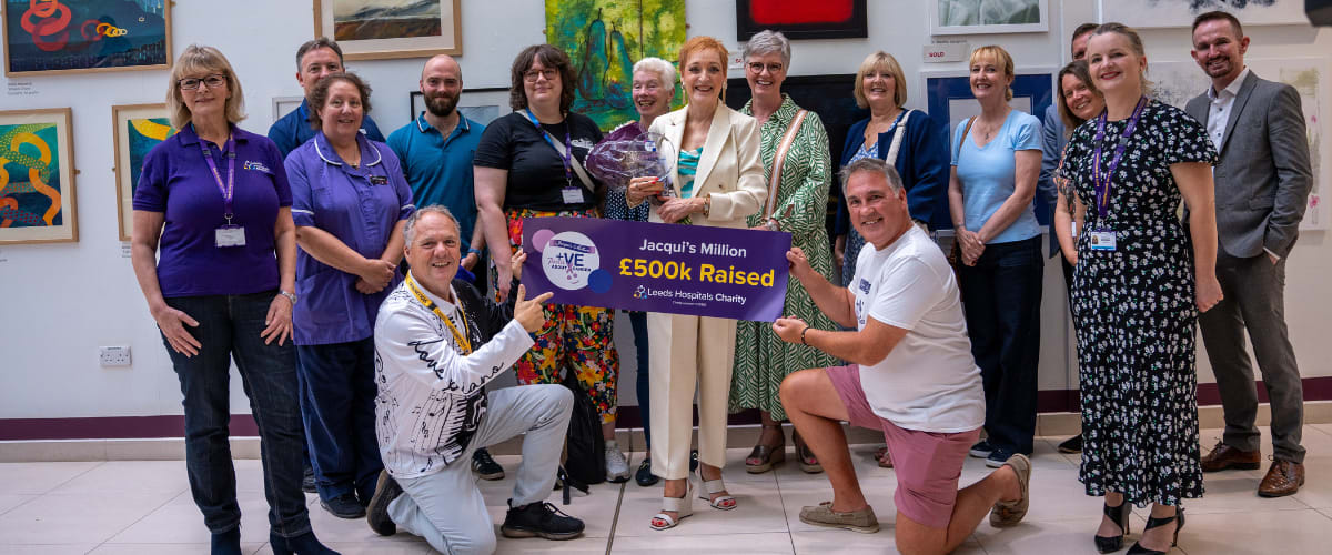 Jacqui Drake with friends, supporters, hospital staff and charity staff at a celebration for her fundraising milestone, holding a banner