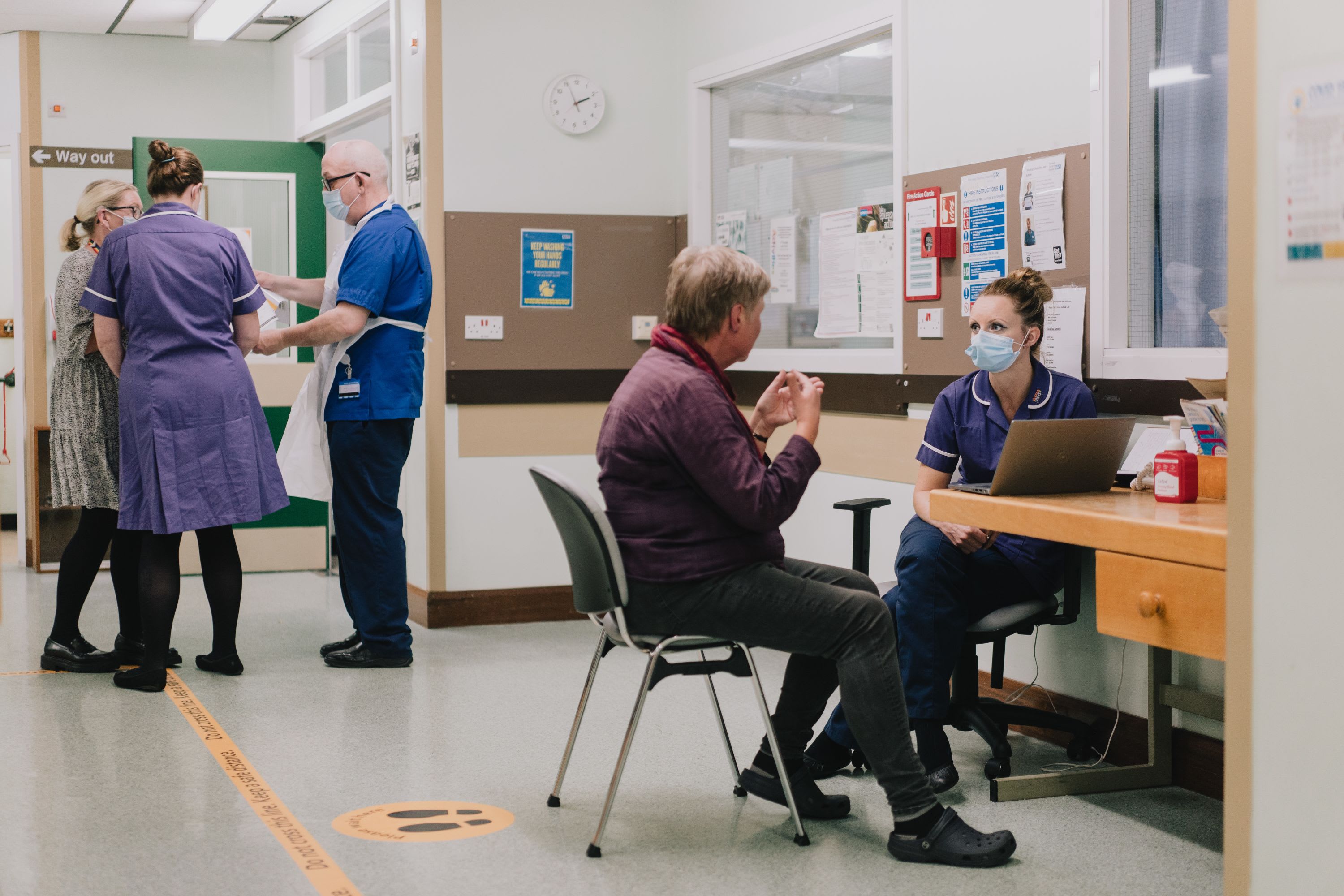 A group of MND staff talk in a small corridor next to an MND patient and clinical nurse sat talking together
