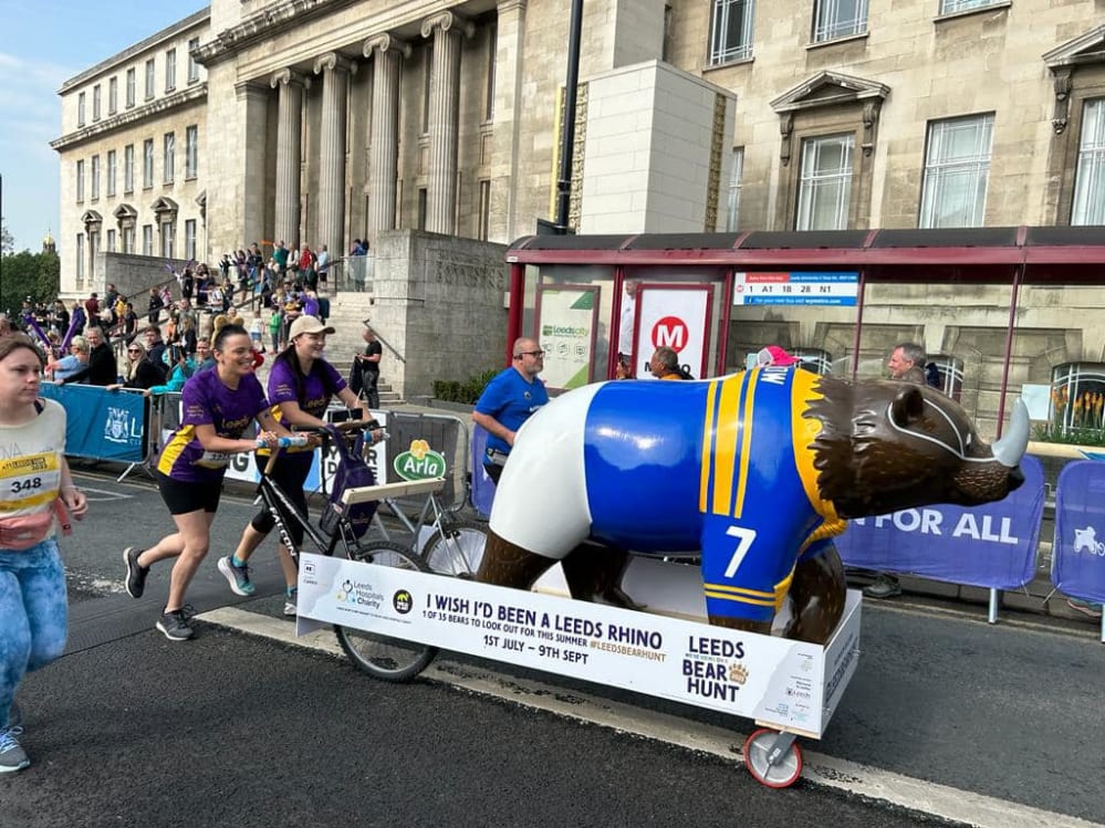 Ducky and Louise from Leeds Hospitals Charity pushing the "I Wish I was a Leeds Rhino" bear around the 2023 Leeds 10k to promote the Leeds Bear Hunt.